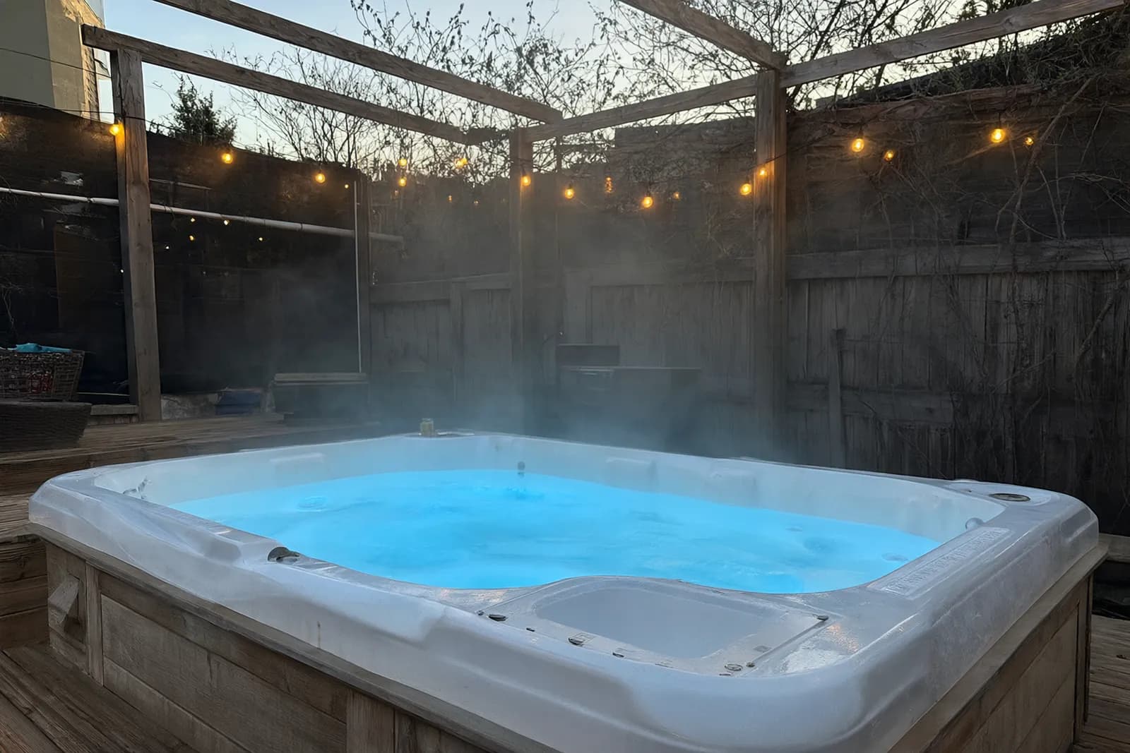 An outdoor hot tub in a wooden deck at dusk, steam rising under string lights